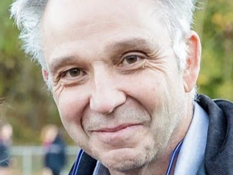 Smiling man with short hair wearing a dark jacket and light shirt in an outdoor setting.