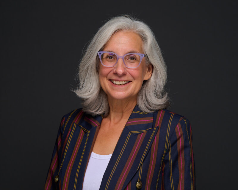 A woman with glasses and gray hair smiles while wearing a striped blazer over a white top.
