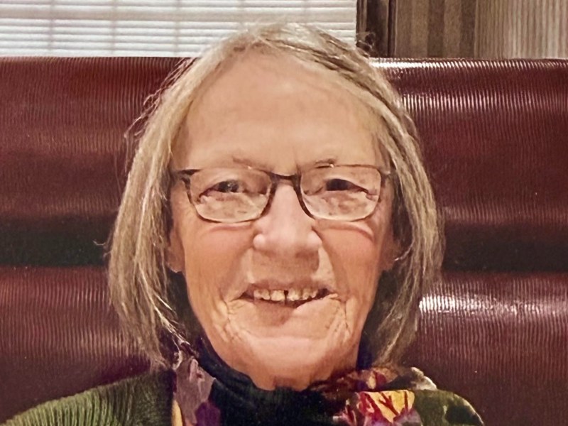 A person with glasses and gray hair smiles while sitting in front of a window. They are wearing a colorful scarf and a green top.
