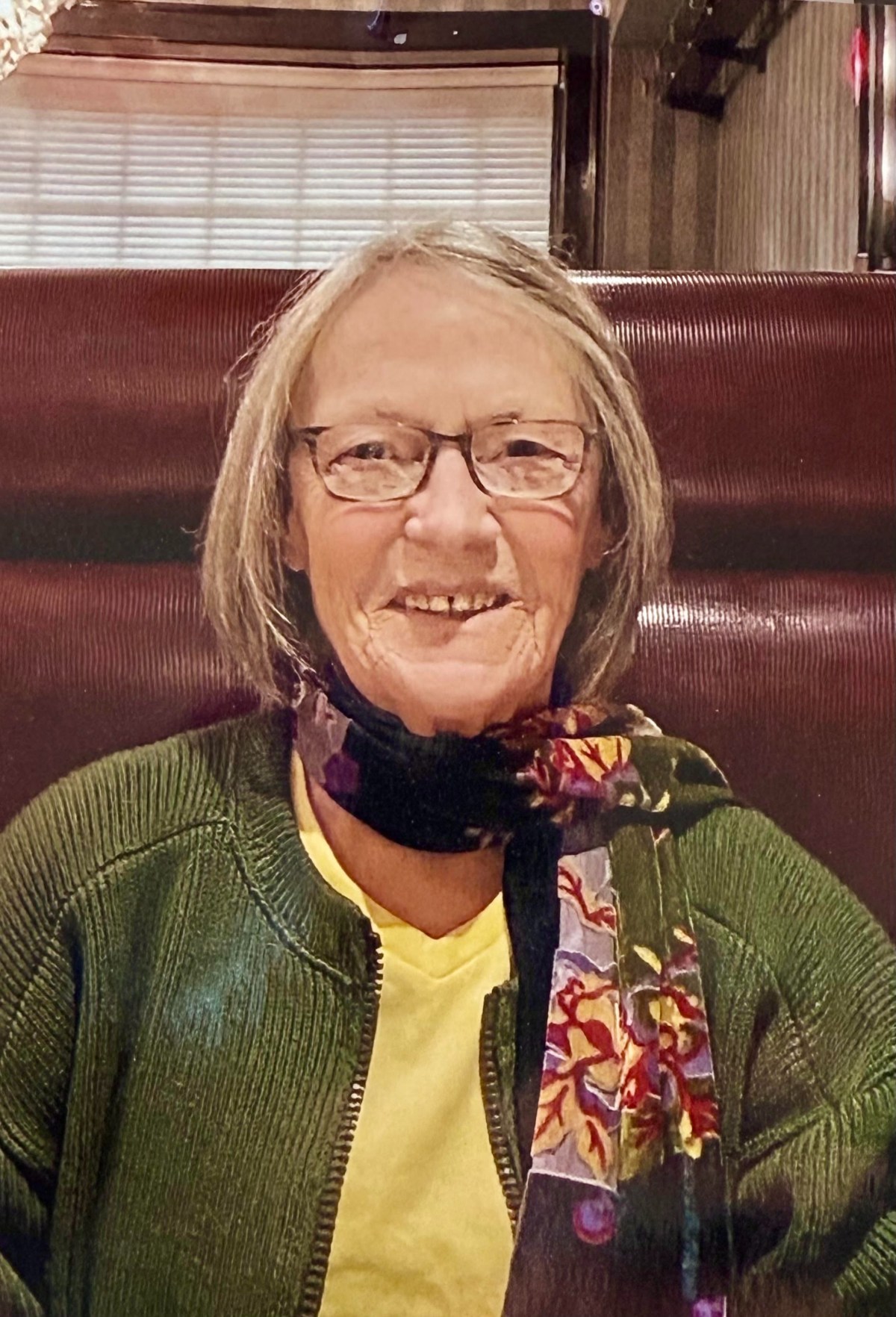 Person with glasses, gray hair, wearing a green sweater and patterned scarf, sitting in a booth and smiling at the camera.