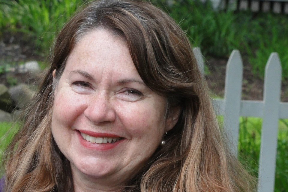 Smiling woman with long brown hair in front of a white picket fence and greenery.
