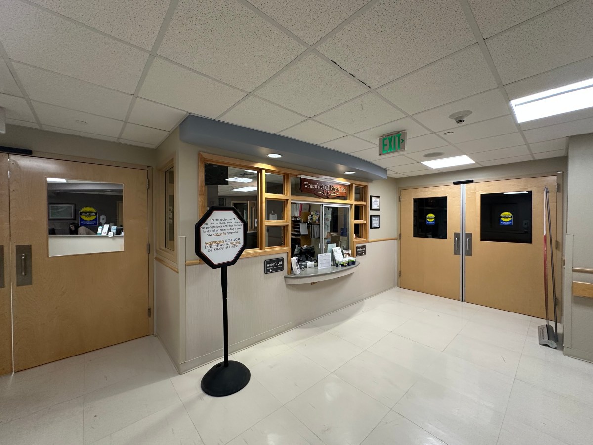 Hospital hallway with a reception desk and decorative sign. Doors labeled with caution tape are visible on the right. Ceiling lights illuminate the area.