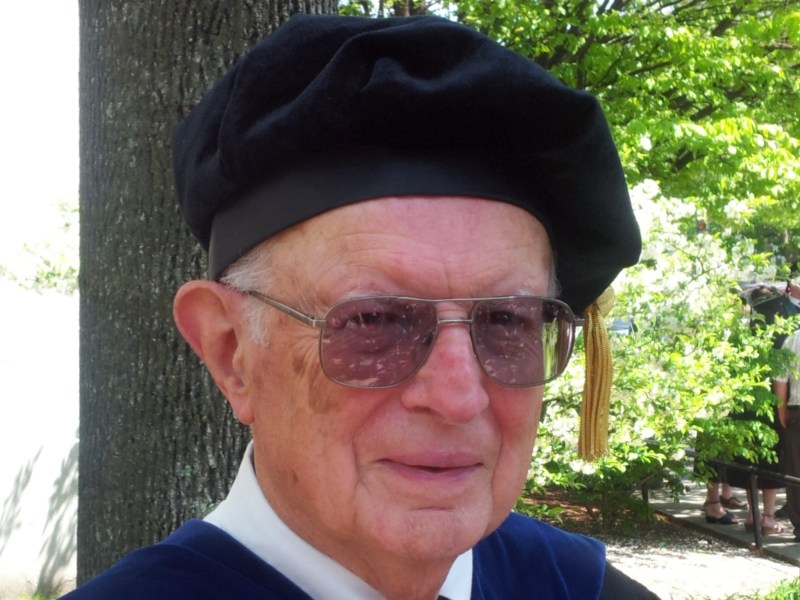 A person wearing graduation attire, including a black cap and tassel, stands outdoors with green foliage in the background.