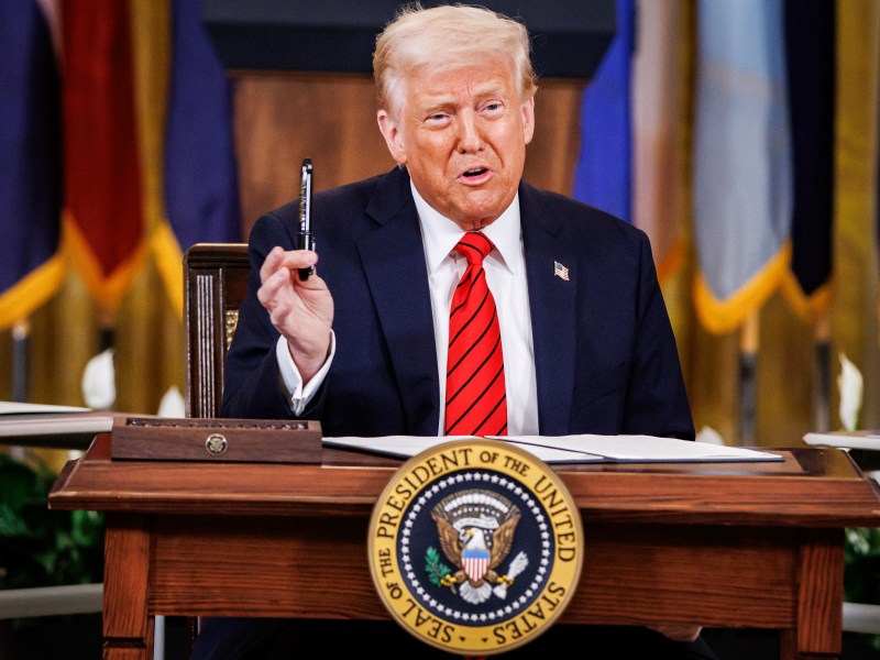 A man in a suit and red tie sits at a desk, holding a black pen, with a presidential seal visible on the desk. Flags are in the background.