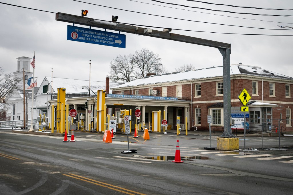 Road border crossing with multiple lanes and stop signs, surrounded by traffic cones and a nearby brick building, under a cloudy sky.