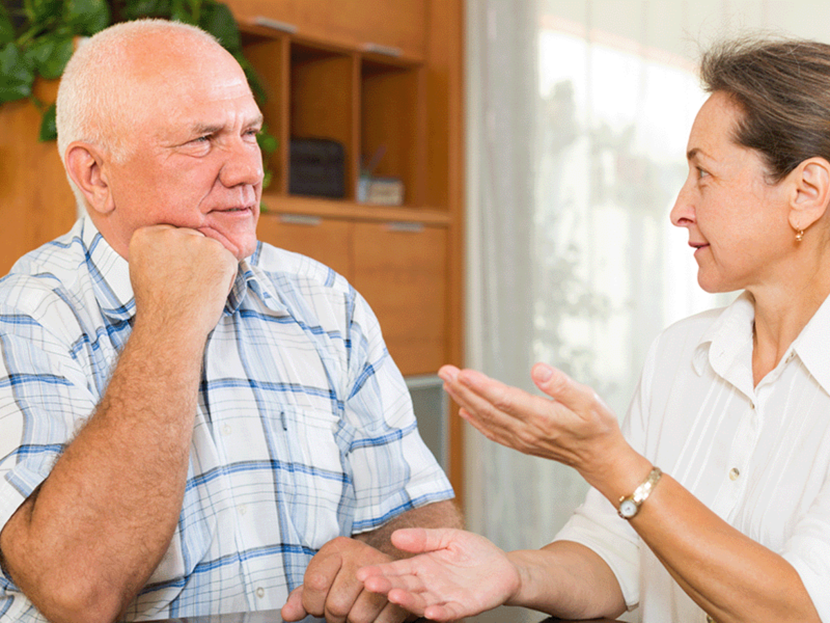 An elderly man and woman sit and converse in a room with wooden shelves in the background. The woman gestures with her hand while the man listens attentively, resting his chin on his hand.