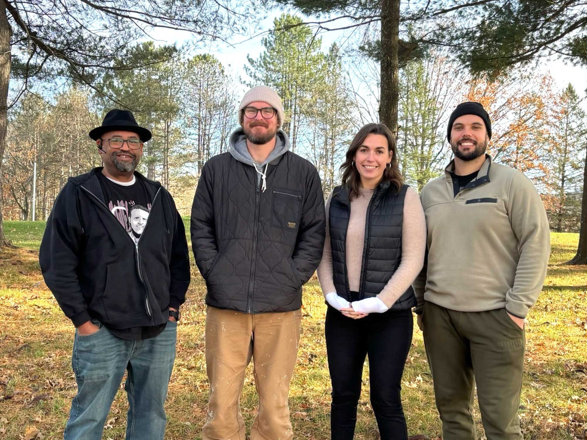 Four people stand outdoors on grass with trees in the background, wearing casual winter clothing and smiling at the camera.