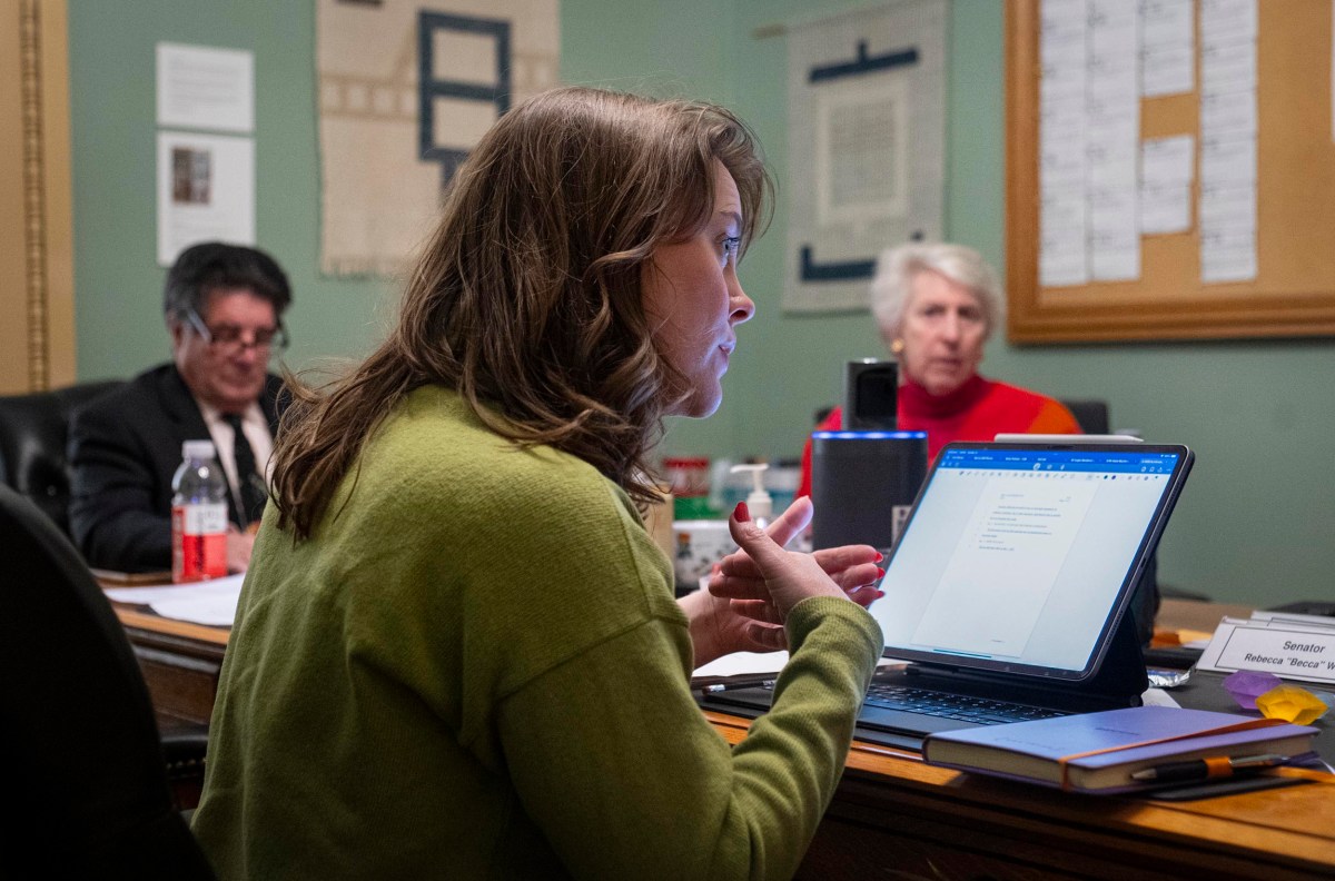 A woman in a green sweater speaks while using a laptop in a meeting room. Two other people sit in the background, one in a red sweater and the other in glasses.