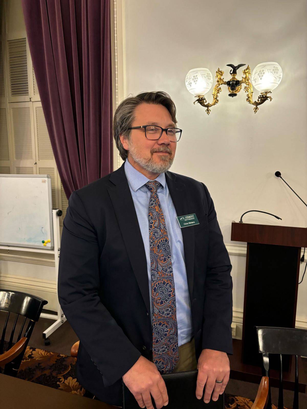 A man in a suit and paisley tie stands in a room with a lectern, whiteboard, and ornate wall sconce.