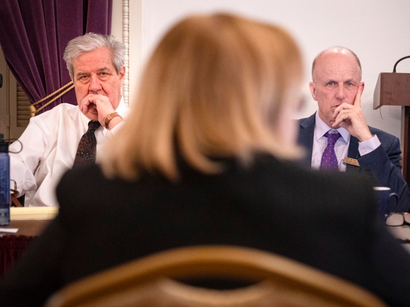 Two men seated at a meeting table appear focused, looking past a person in the foreground. A lectern is visible in the background.