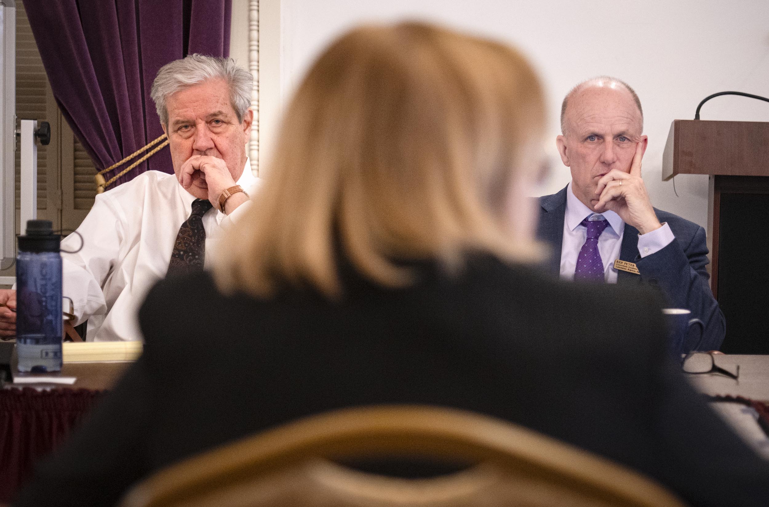 Two men seated at a meeting table appear focused, looking past a person in the foreground. A lectern is visible in the background.