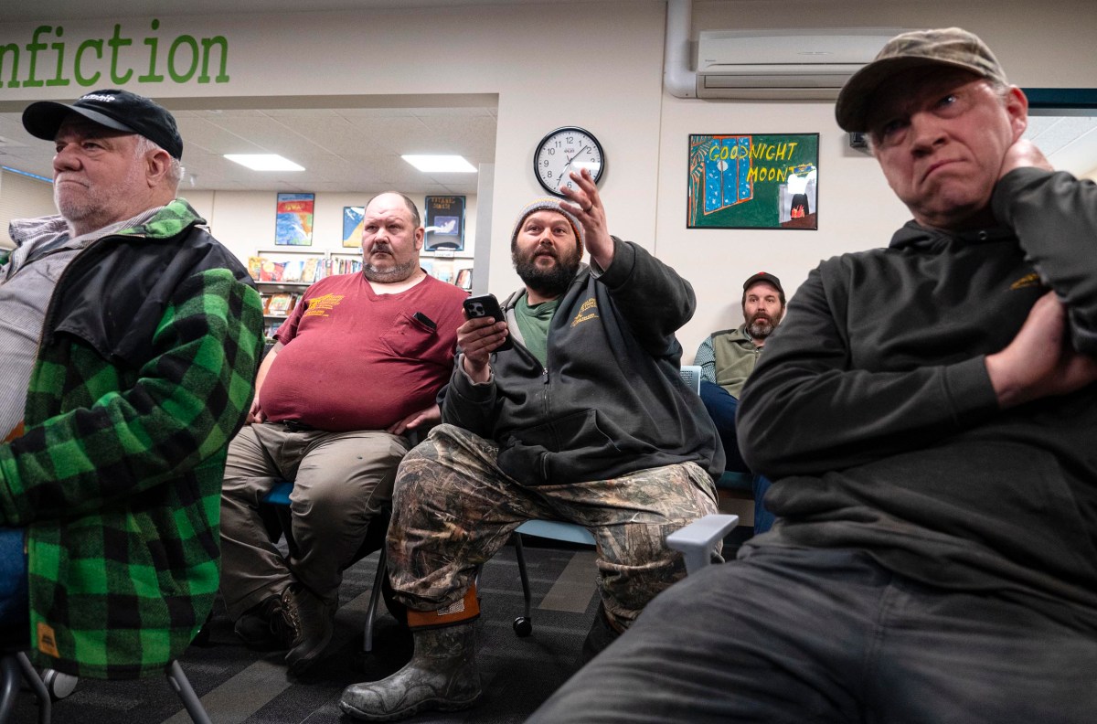 A group of men seated indoors, with one holding a phone and gesturing. Some look attentive while others appear disinterested. The setting includes bookshelves and a clock on the wall.