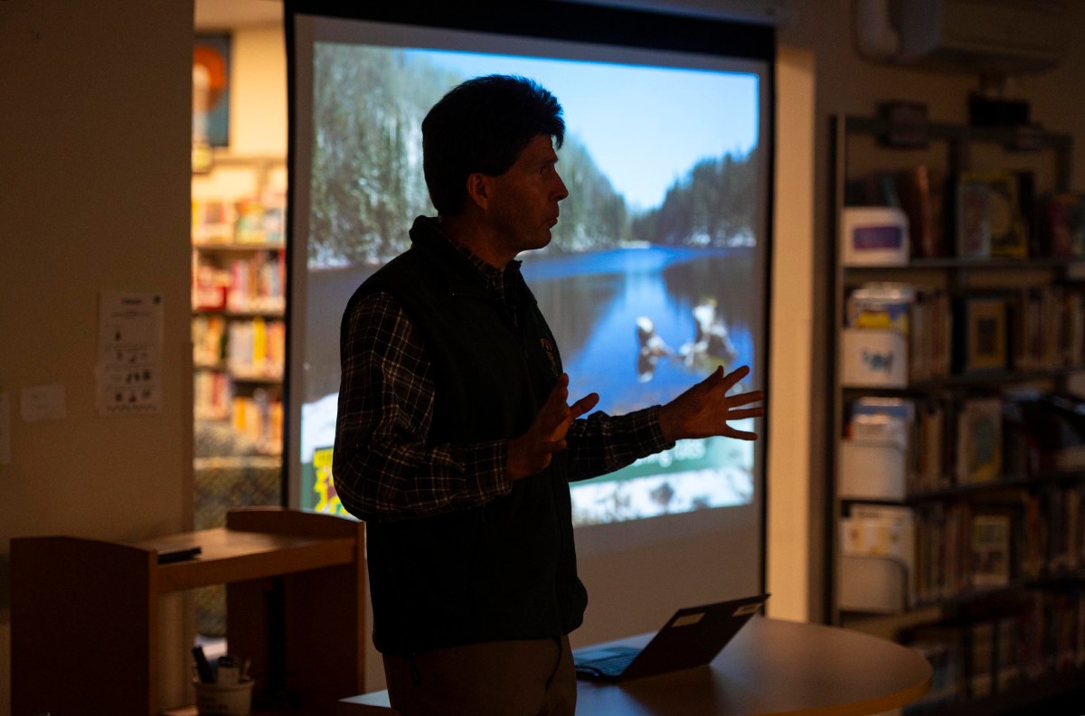 A man gives a presentation in a dimly lit room, gesturing with his hands. A projected image of a lake scene is visible behind him.