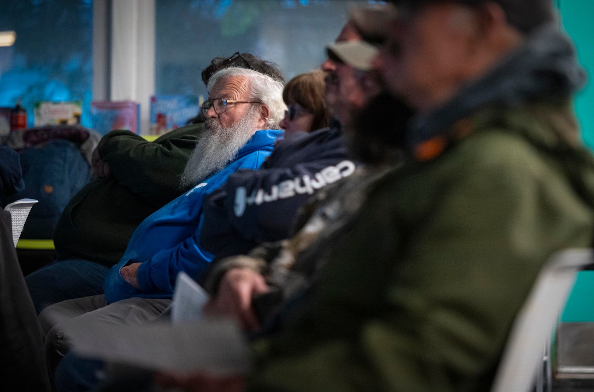 A group of people sit in a room, some wearing hats and jackets. A man with a long white beard wearing a blue hoodie is in focus. They appear to be listening attentively.
