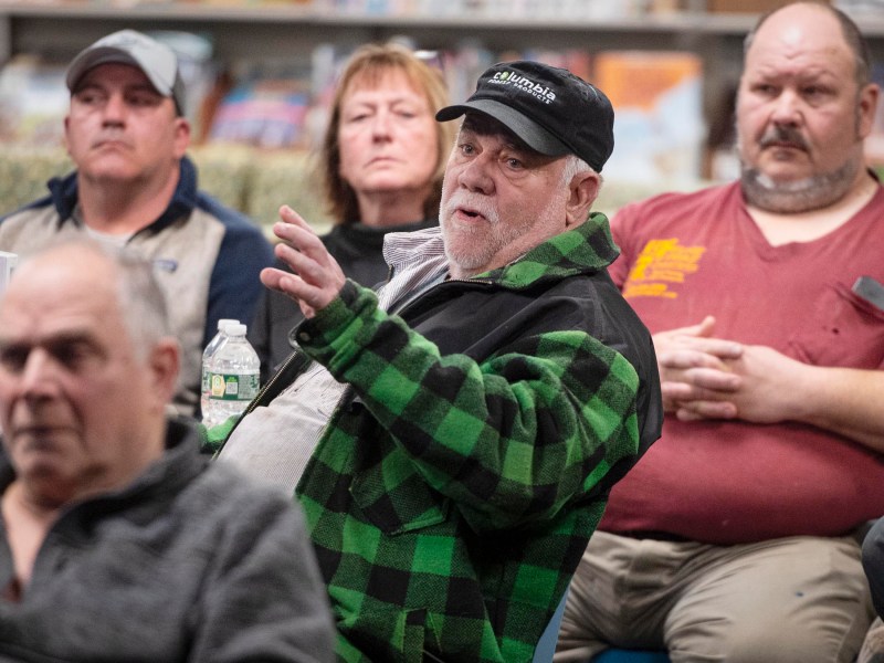 A group of people seated indoors, with one man in a green plaid jacket and black cap speaking, while others listen attentively.