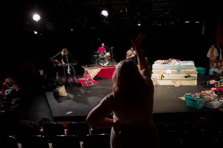 A person stands in a theater audience, raising their arm toward a stage where three musicians are playing among scattered clothing and beds.
