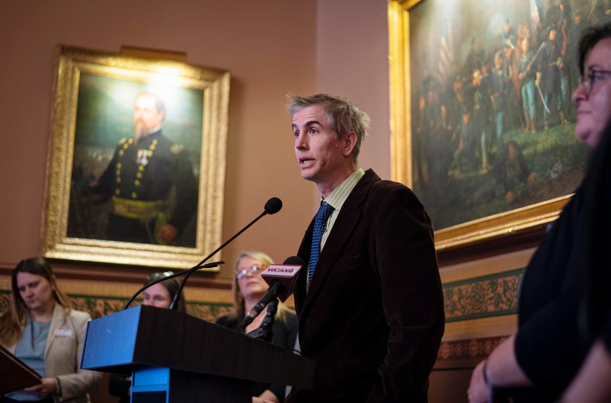 A person speaking at a podium, surrounded by others, with framed paintings in the background.
