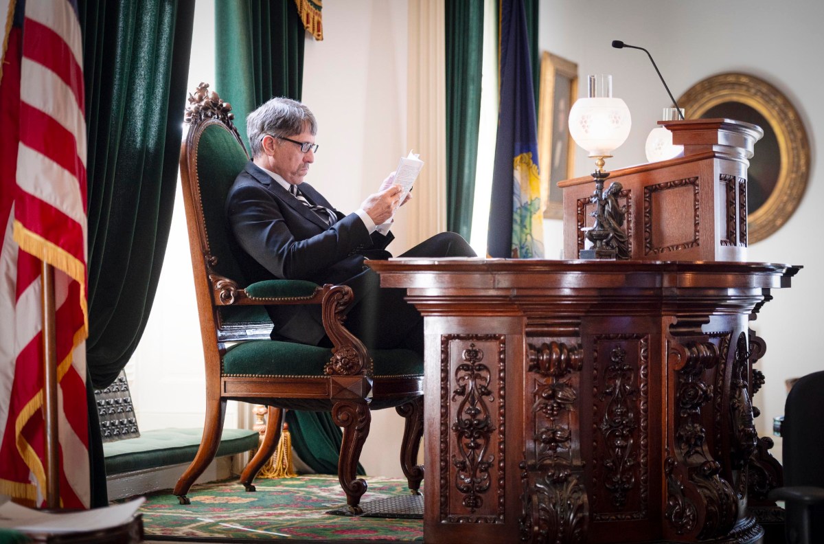 Man sitting on an ornate chair reading papers at a wooden desk in a room with heavy curtains and an American flag.