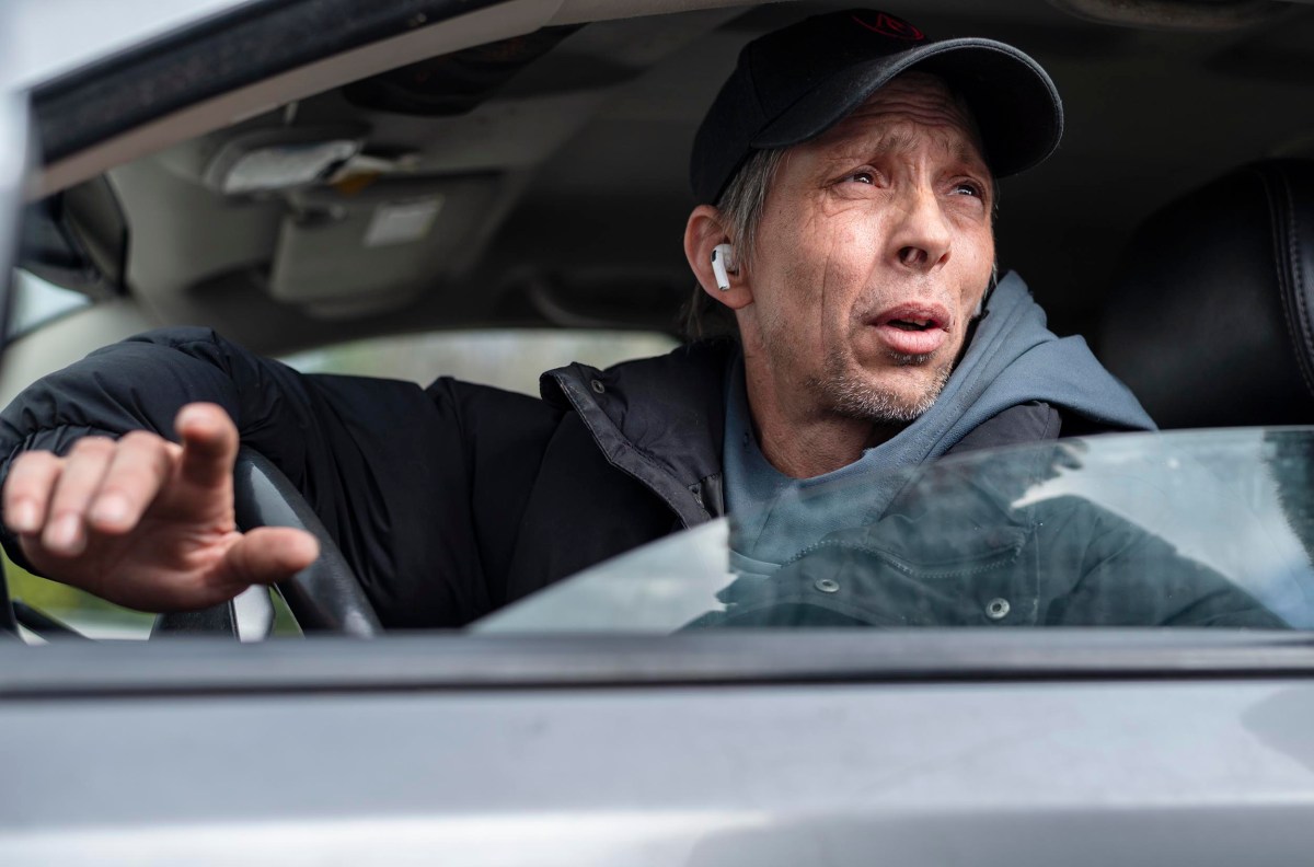 Person wearing a cap and earbuds, sitting inside a car with the window down, gesturing with a hand.