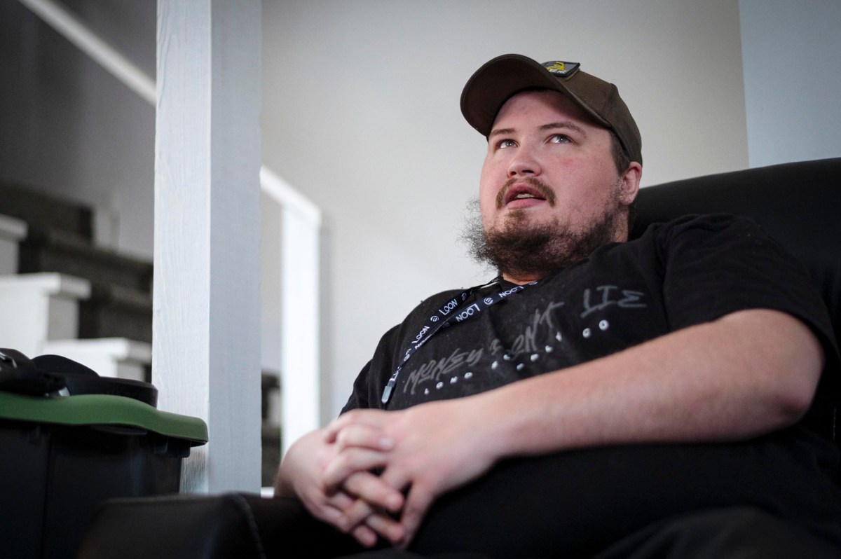 A person with a beard and wearing a cap sits in a chair, hands folded. Steps and a white wall are in the background.