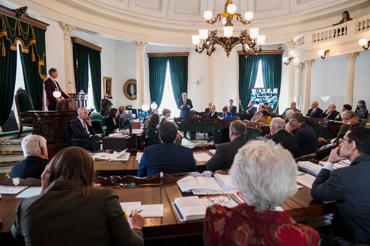 A formal meeting in a large room with people seated around tables, listening to a speaker. There's ornate decor, high ceilings, and a prominent light fixture.