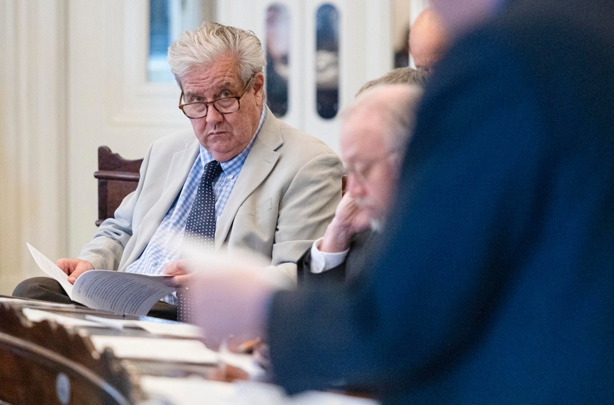 An older man in a light suit sits at a table looking at papers, surrounded by others in a formal setting.