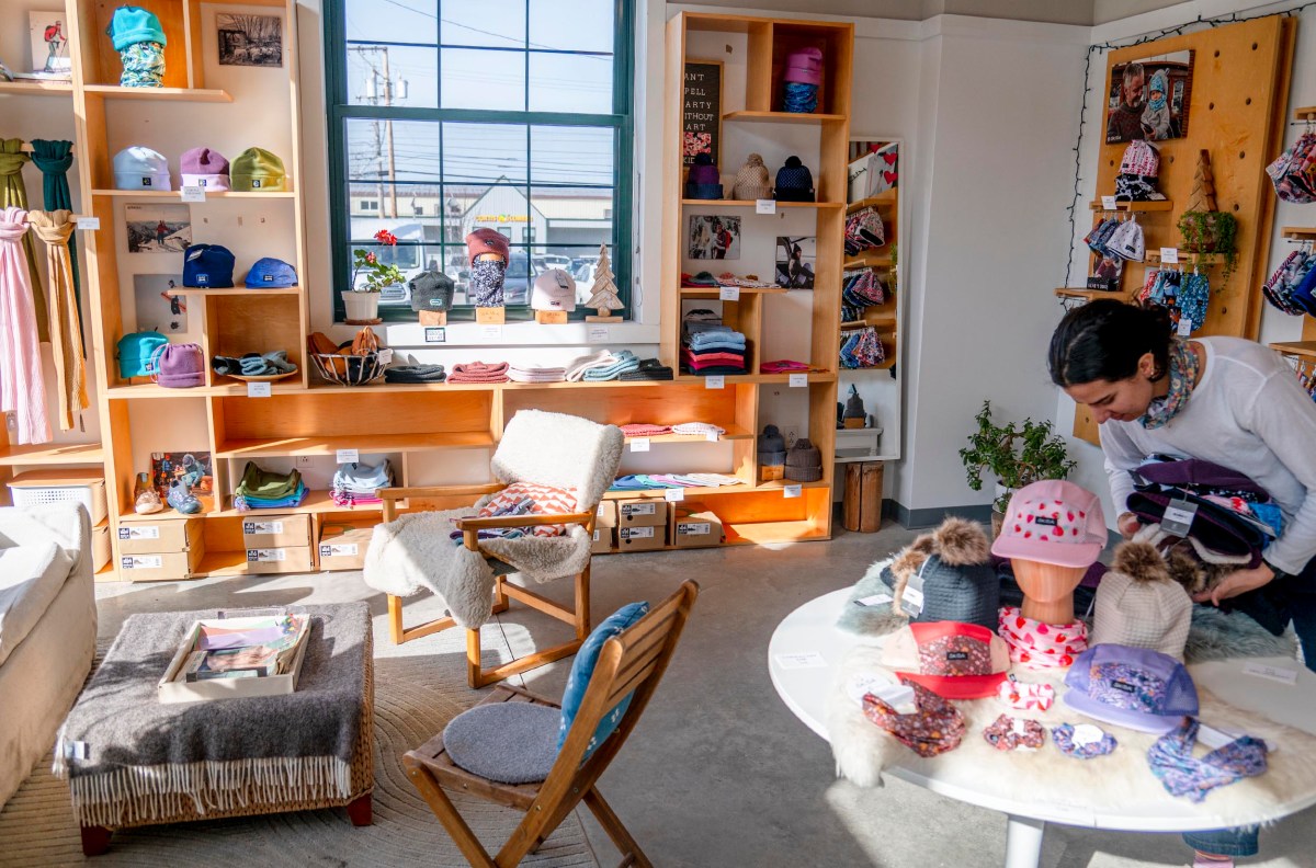 A person browses hats and clothing in a well-lit store with wooden shelves, a window, and a circular display table. Various hats are arranged on mannequins and shelves.