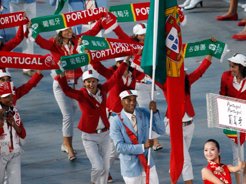 Portuguese athletes in red and white uniforms march with flags and scarves labeled “Portugal” during an Olympic opening ceremony. A woman leads with a sign showing Portugal in English and Chinese.