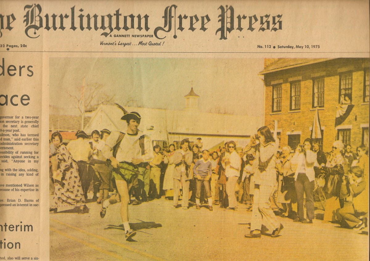 A historical parade features a person in a colonial costume leading a procession on a sunny day, surrounded by onlookers and a brick building in the background.