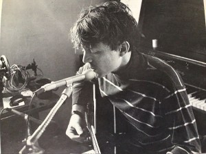 Person with tousled hair plays guitar and sings into a microphone near a piano in a dimly lit room. Black and white photo.