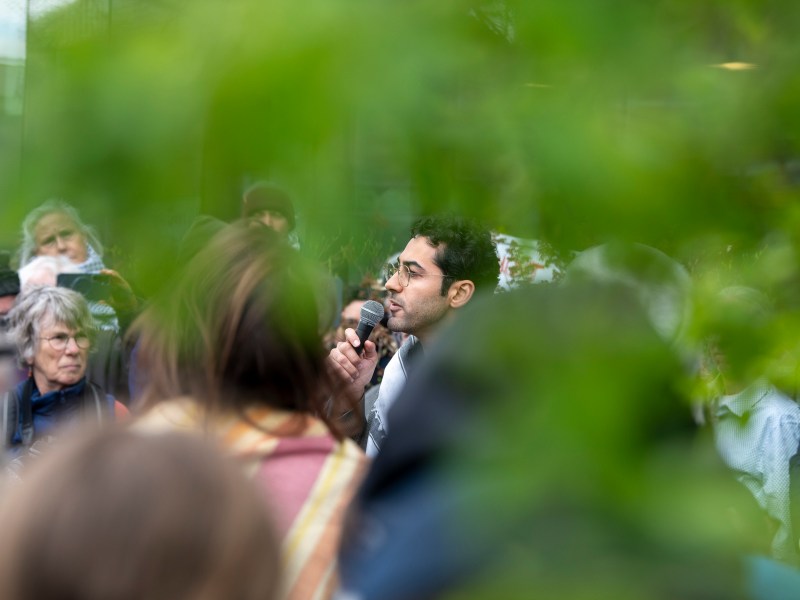 A man speaks into a microphone at an outdoor gathering, partially obscured by green foliage, with people listening in the background.
