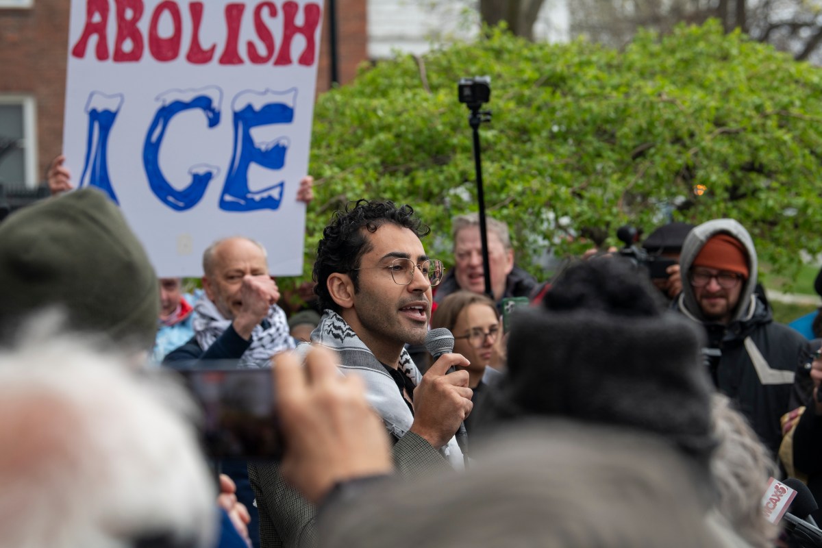 A man speaks into a microphone at an outdoor protest, with people holding signs and a prominent "Abolish ICE" sign visible in the background.