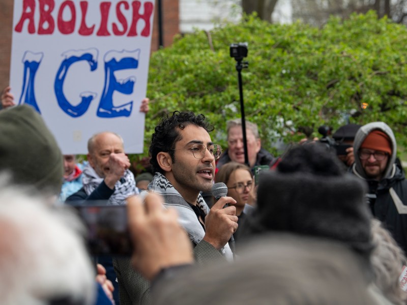 A man speaks into a microphone at an outdoor protest, with people holding signs and a prominent "Abolish ICE" sign visible in the background.