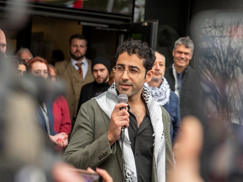 A man in glasses speaks into a microphone outside a building, surrounded by a group of people, some of whom are watching him attentively.