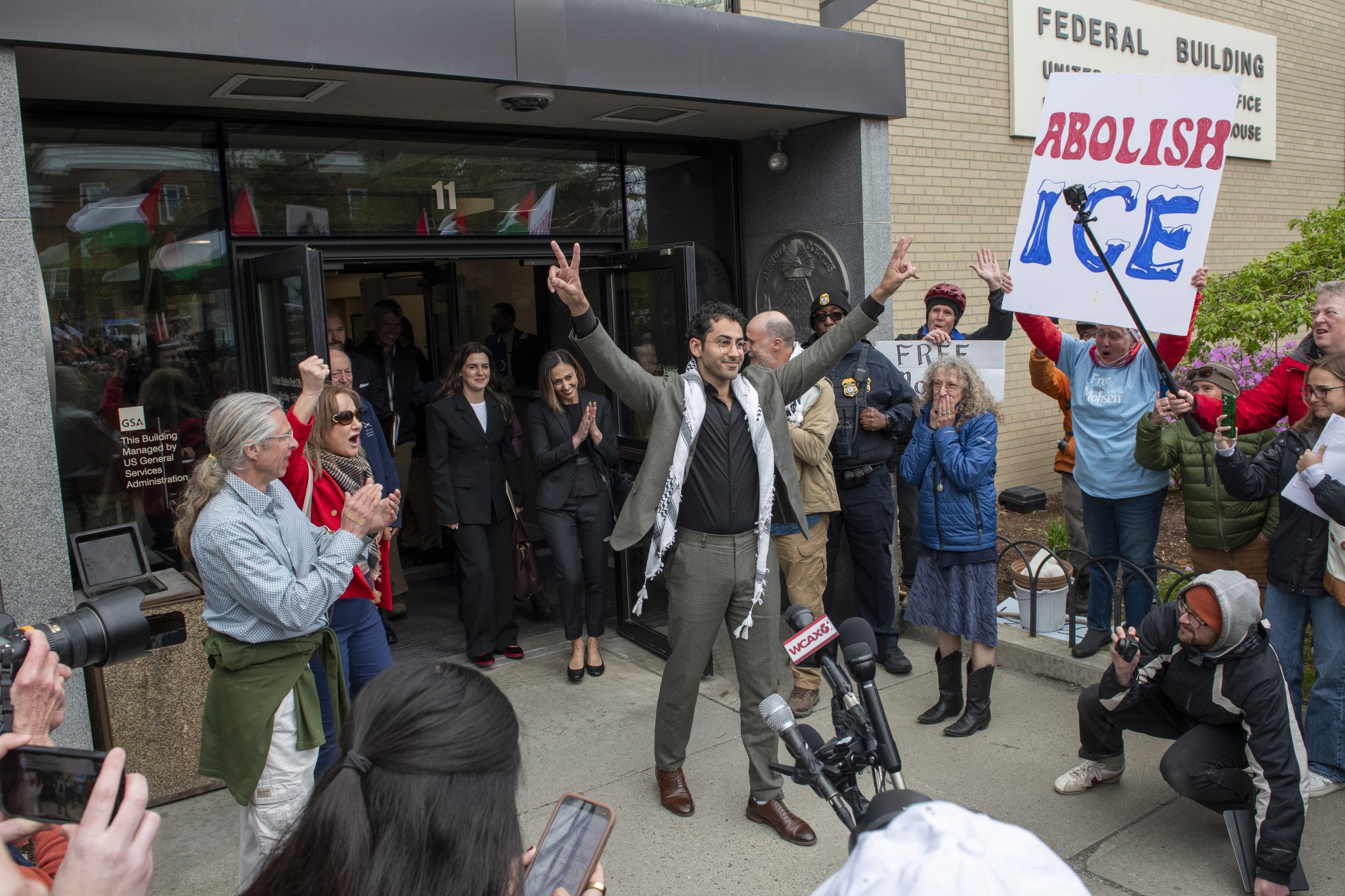 A man stands outside a federal building with raised arms, surrounded by cheering supporters and a visible "Abolish ICE" sign.