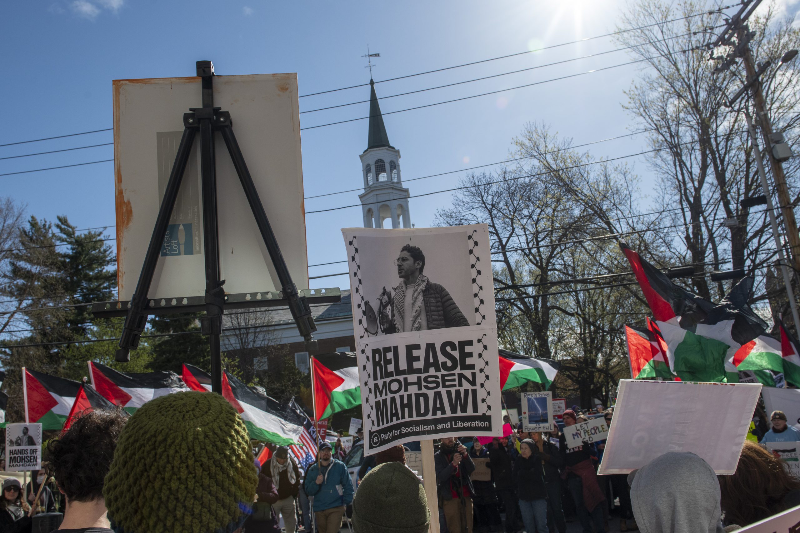 A crowd holds flags and protest signs, including one reading "Release Mohsen Mahdawi," with a church steeple and power lines visible in the background.