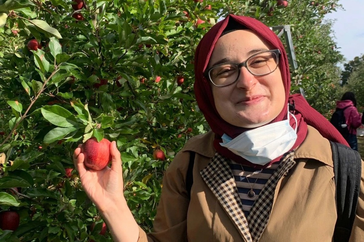 A person in glasses and a headscarf holds a red apple in an orchard, with a partially lowered mask around their neck. Others are visible in the background among green foliage.