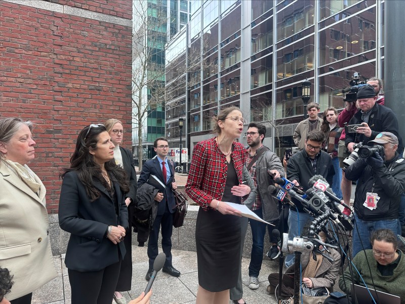 A woman speaks at a press conference outdoors, surrounded by several people and journalists with cameras and microphones. Brick and glass buildings are in the background.