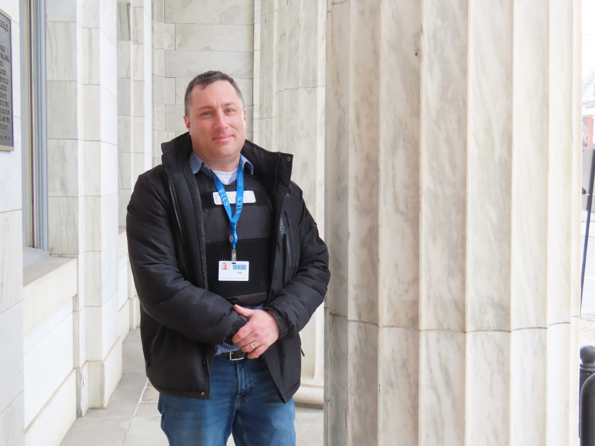 A man in a black jacket and jeans stands beside a marble column in an outdoor hallway, wearing a name badge and lanyard.