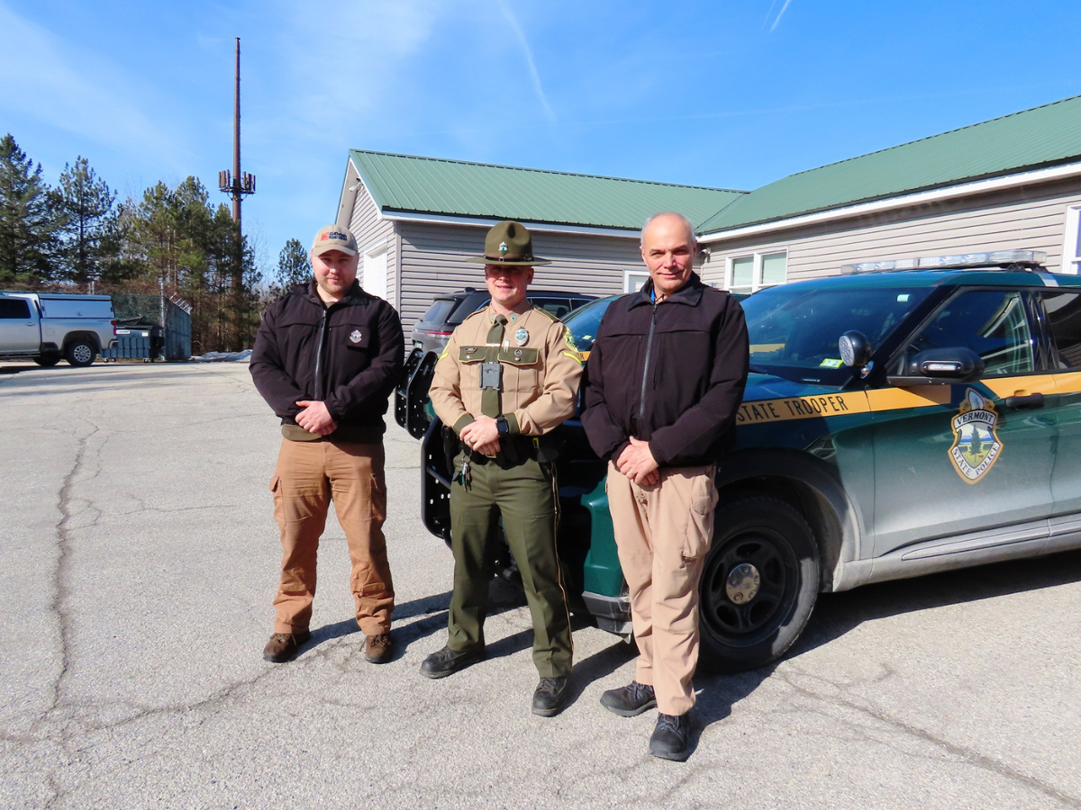 Three law enforcement officers stand in front of a patrol car outside a building on a sunny day.