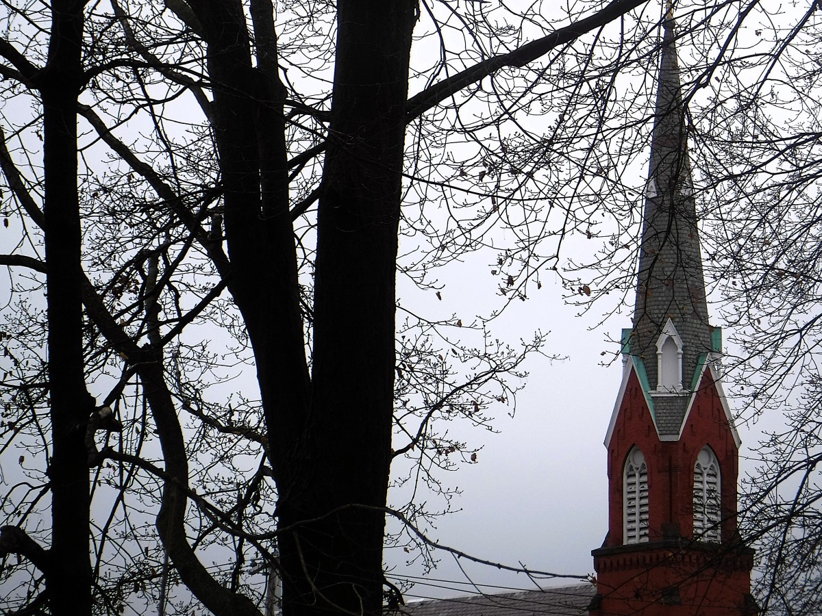 Red brick church steeple with white trim and pointed roof, seen behind leafless trees on an overcast day.