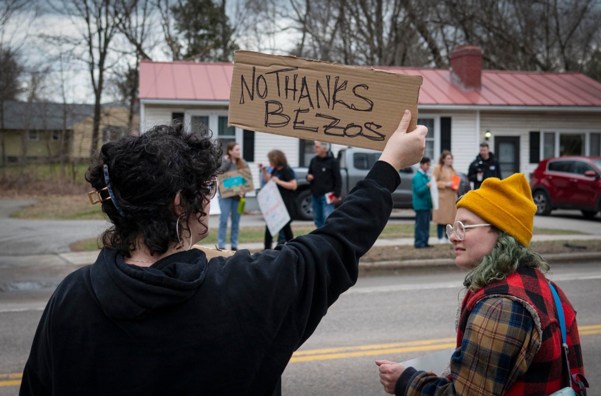 Person holds a sign saying "No Thanks Bezos" at a protest, with others in the background holding signs on a residential street.