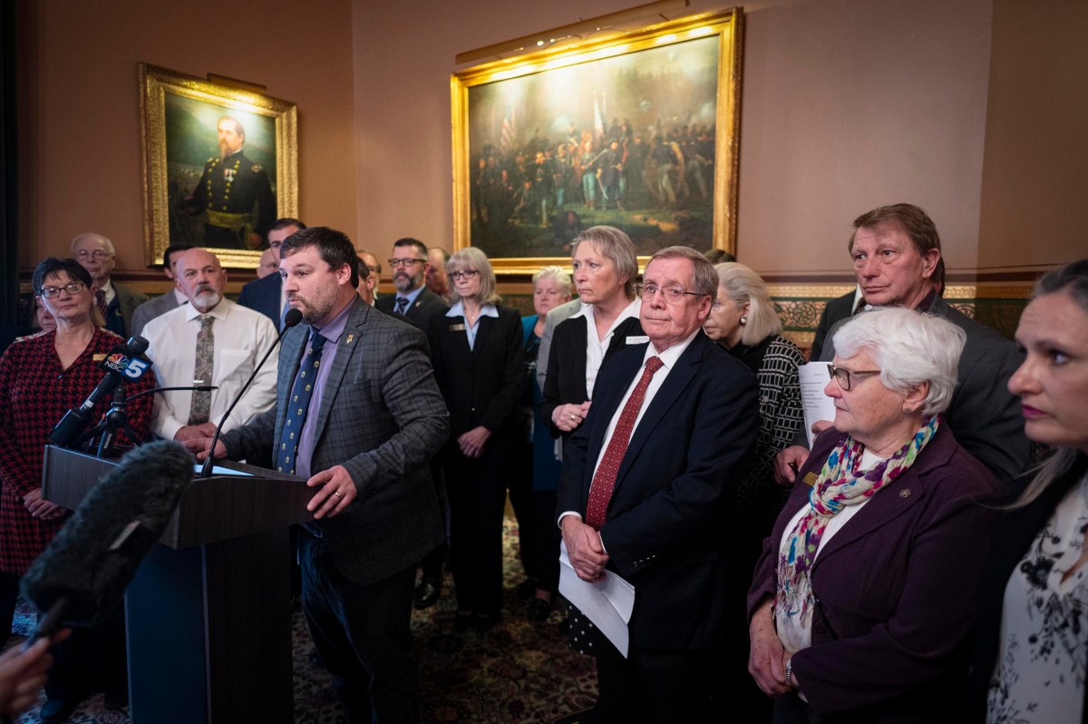 A group of people stands in a room with ornate paintings. A man at a podium addresses microphones, while others listen attentively.