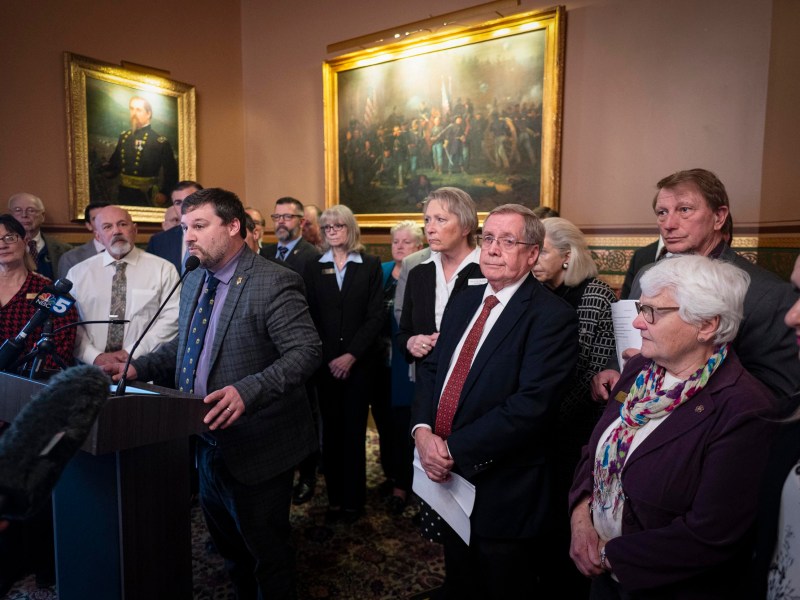 A group of people stands in a room with ornate paintings. A man at a podium addresses microphones, while others listen attentively.