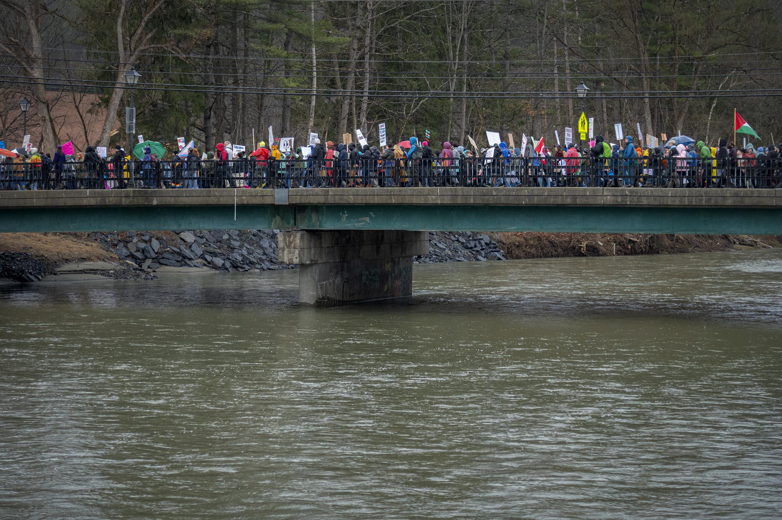 A large group of people holding signs is walking across a bridge over a wide river on a cloudy day.