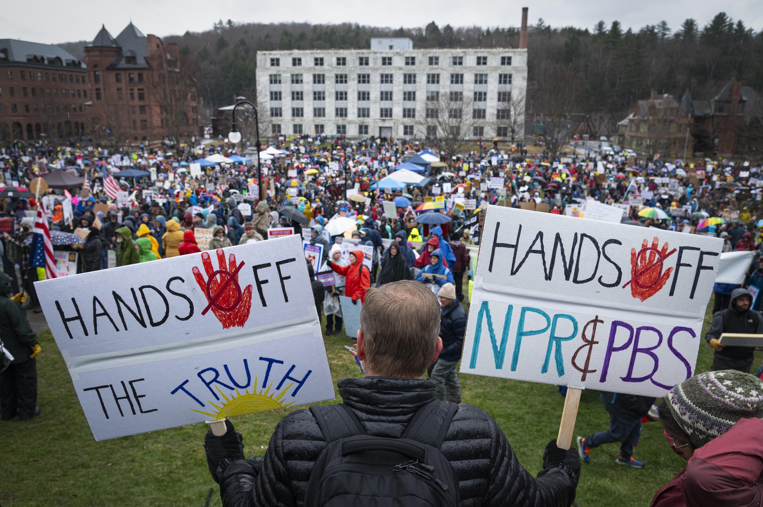 Crowd gathered at a protest. Person in foreground holds signs reading "Hands Off The Truth" and "Hands Off NPR & PBS.