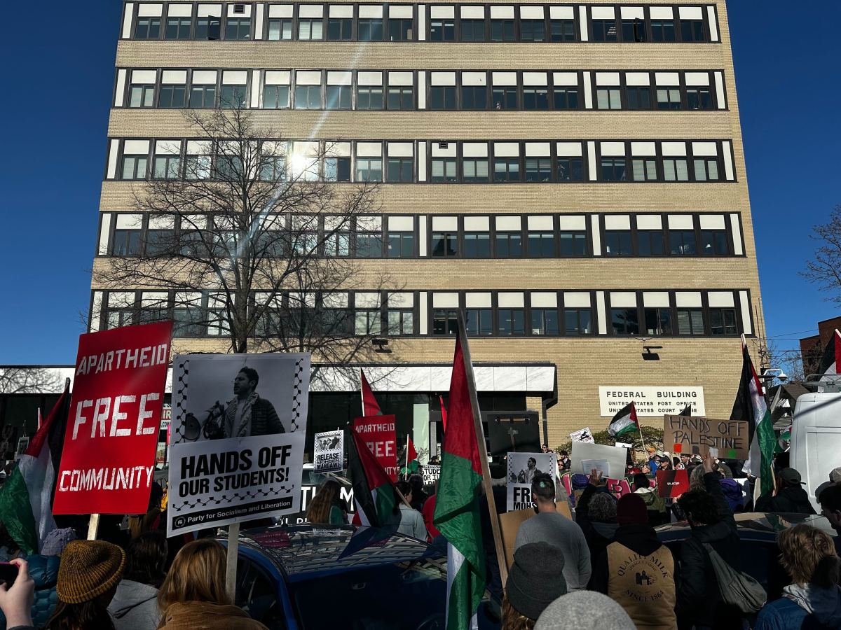 A crowd of protesters stands outside a multistory building, holding signs with messages including "Free Palestine" and "Hands Off Our Students.