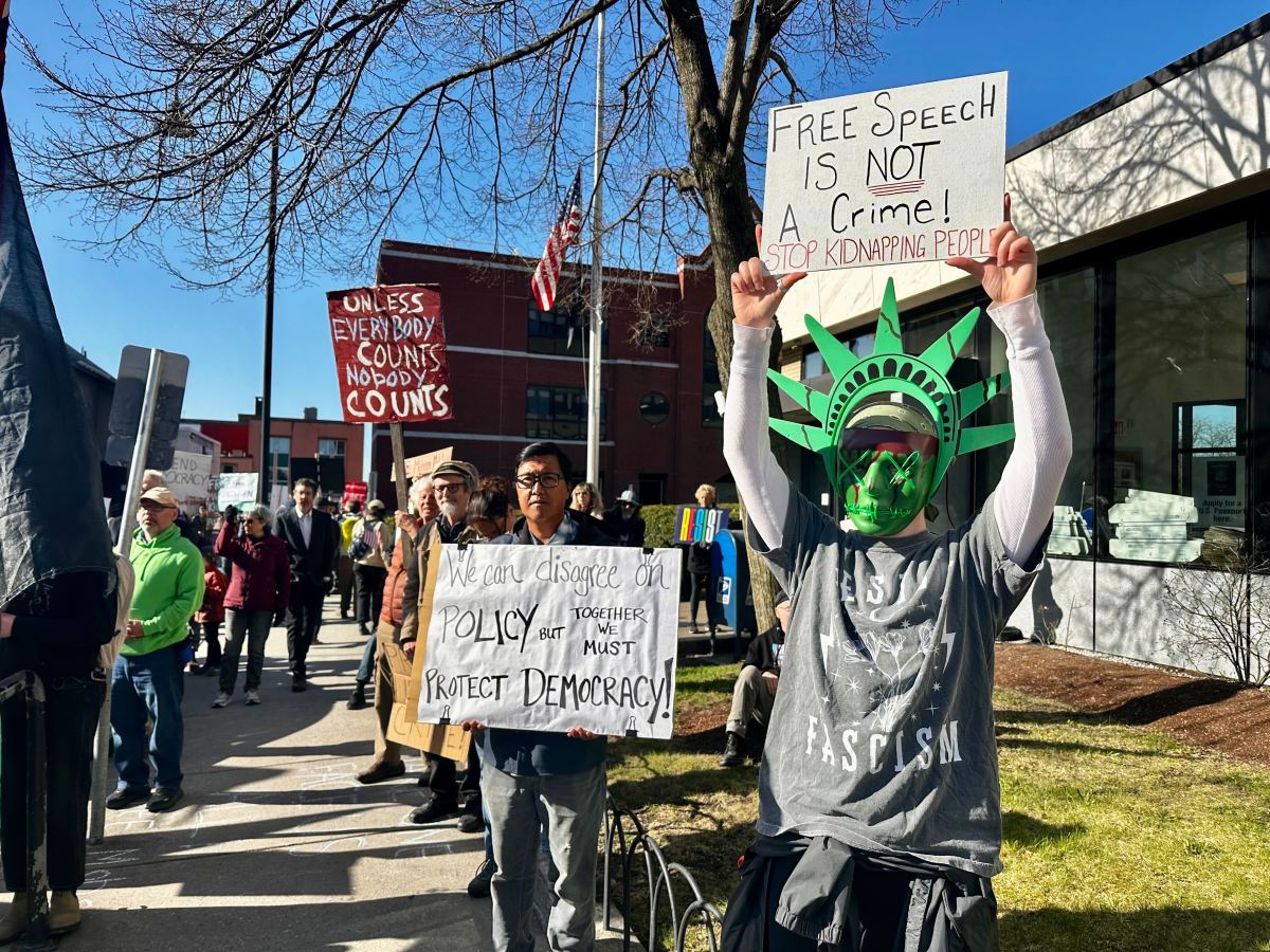 A group of protesters march outside, holding signs about free speech, democracy, and policy. One person wears a Statue of Liberty crown and mask.
