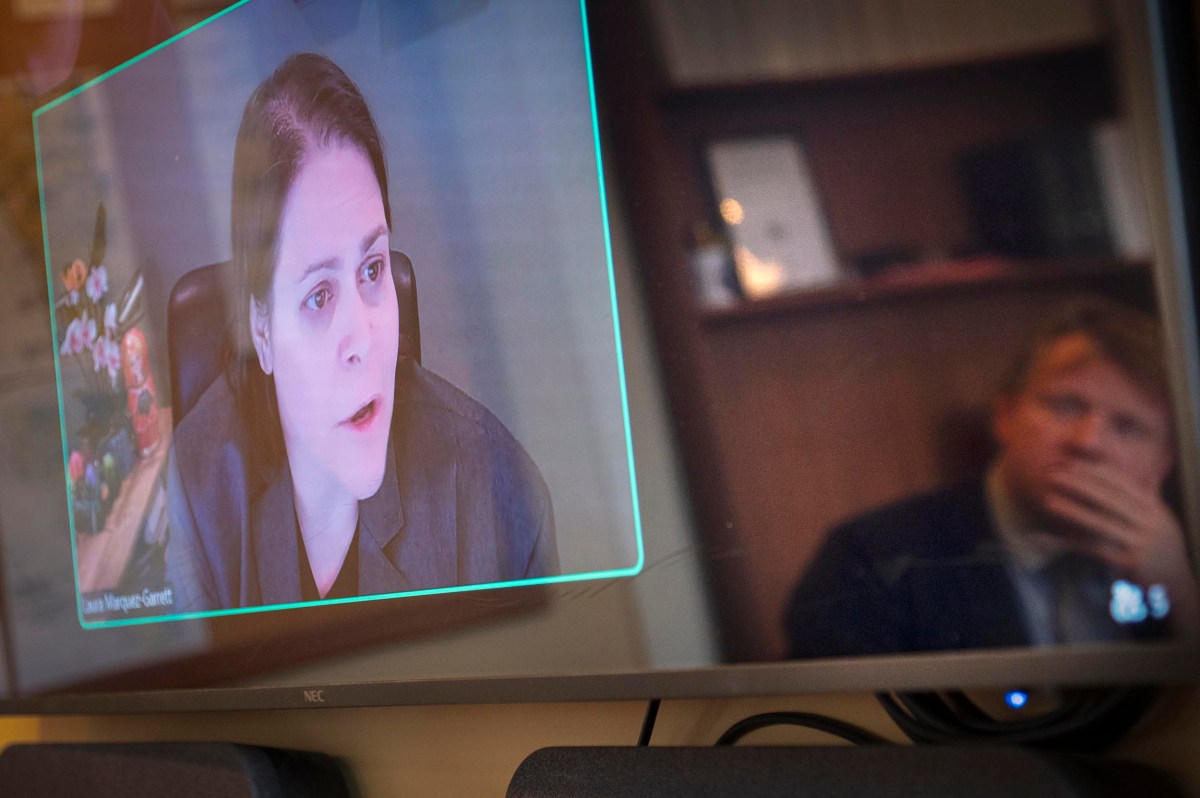 A person in a video call is displayed on a screen while another person is seen in reflection, watching attentively.