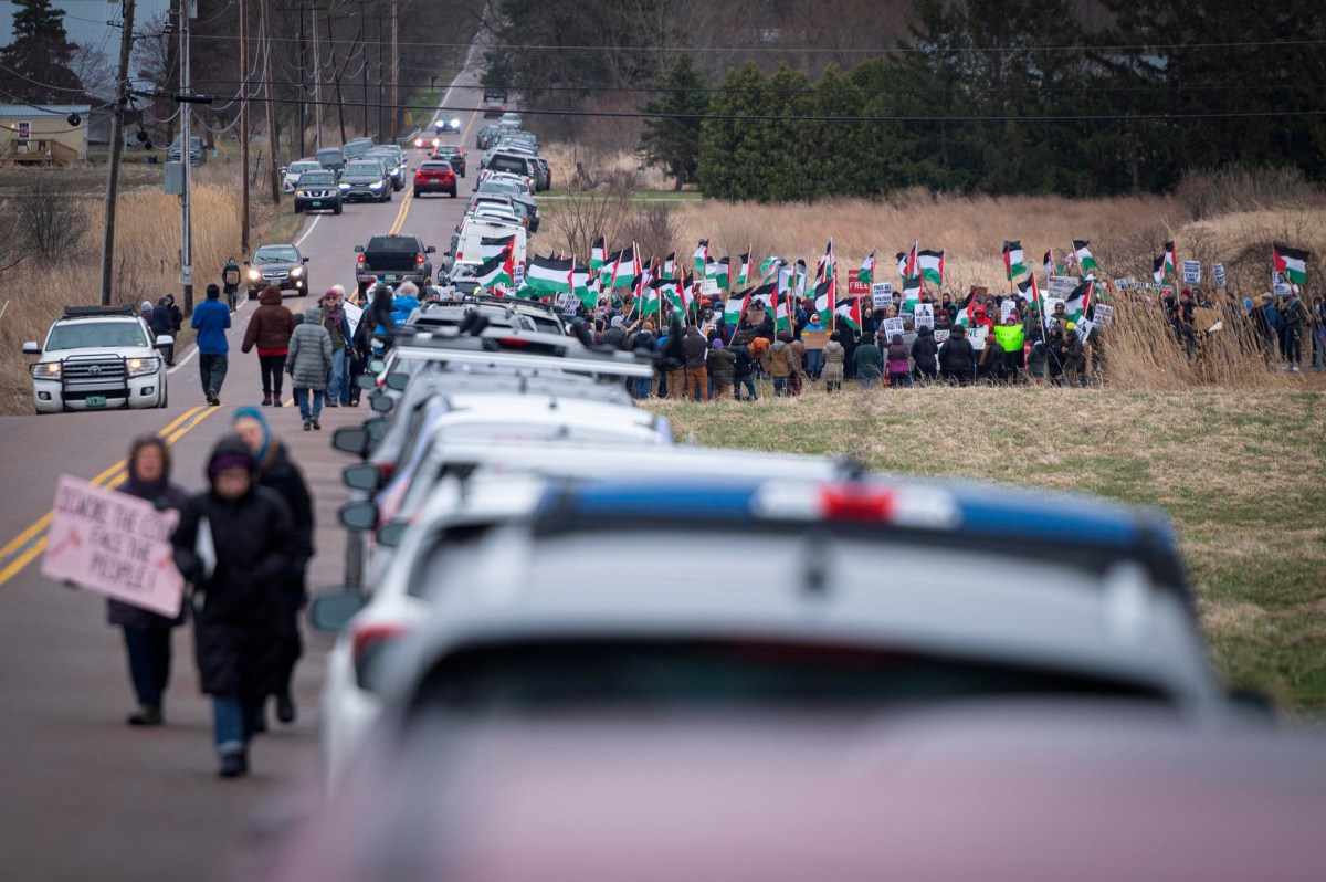 A line of parked cars along a rural road with a group of people holding signs and flags gathered in the background; two people walk beside the vehicles.